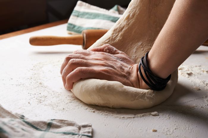 Hands kneading bread dough on a white kitchen surface, with the dough stretching into the air.