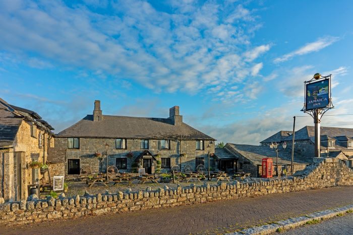 View of the Jamaica Inn from the road, with an old Inn sign and stone wall construction. Blue sky with a cloud just above.