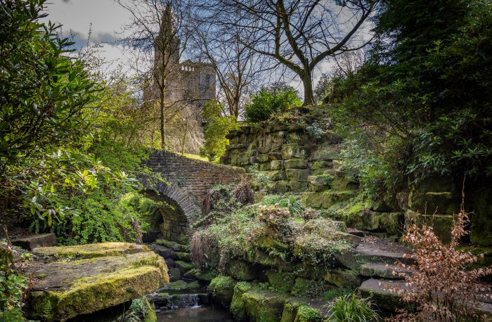 Alt: A stone bridge surrounded by foliage and mossy stone, Dunfermline Abbey in the background.