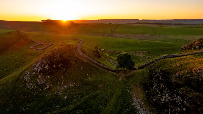 Aerial view of Hadrian’s Wall at Sycamore Gap, with the sun setting over green grassland. Roman fort ruins visible along the path.