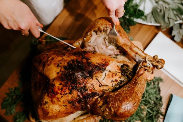 A person cutting a roasted turkey on a bed of herbs and chopping board