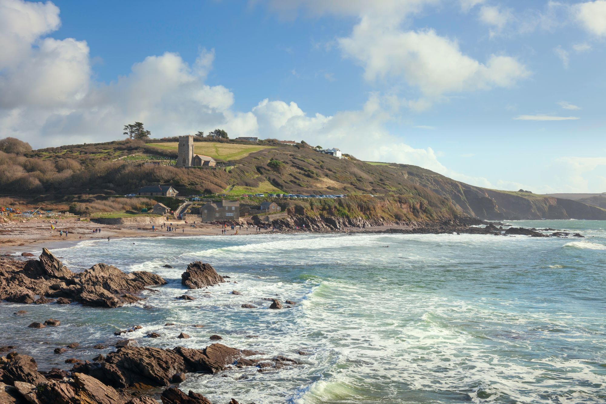 View of Wembury Beach from the rocky shore, with waves crashing on the rocks and sand. A church sits on the cliff in the background.